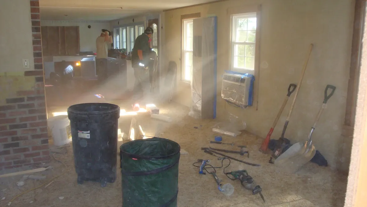 Interior mid-demolition: plaster dust hanging in the light, brooms and buckets beside a brick chimney breast.