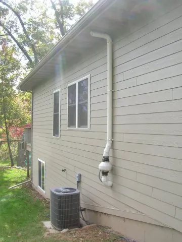 A white PVC radon discharge stack strapped up the siding of a house with an inline fan housing, terminating above the eave.